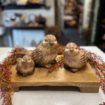 Three hand-carved wooden bird figurines in natural brown tones, arranged on a rectangular wooden base with dried autumn berries in a blurred indoor setting.