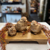 Three hand-carved wooden bird figurines in natural brown tones, arranged on a rectangular wooden base with dried autumn berries in a blurred indoor setting.