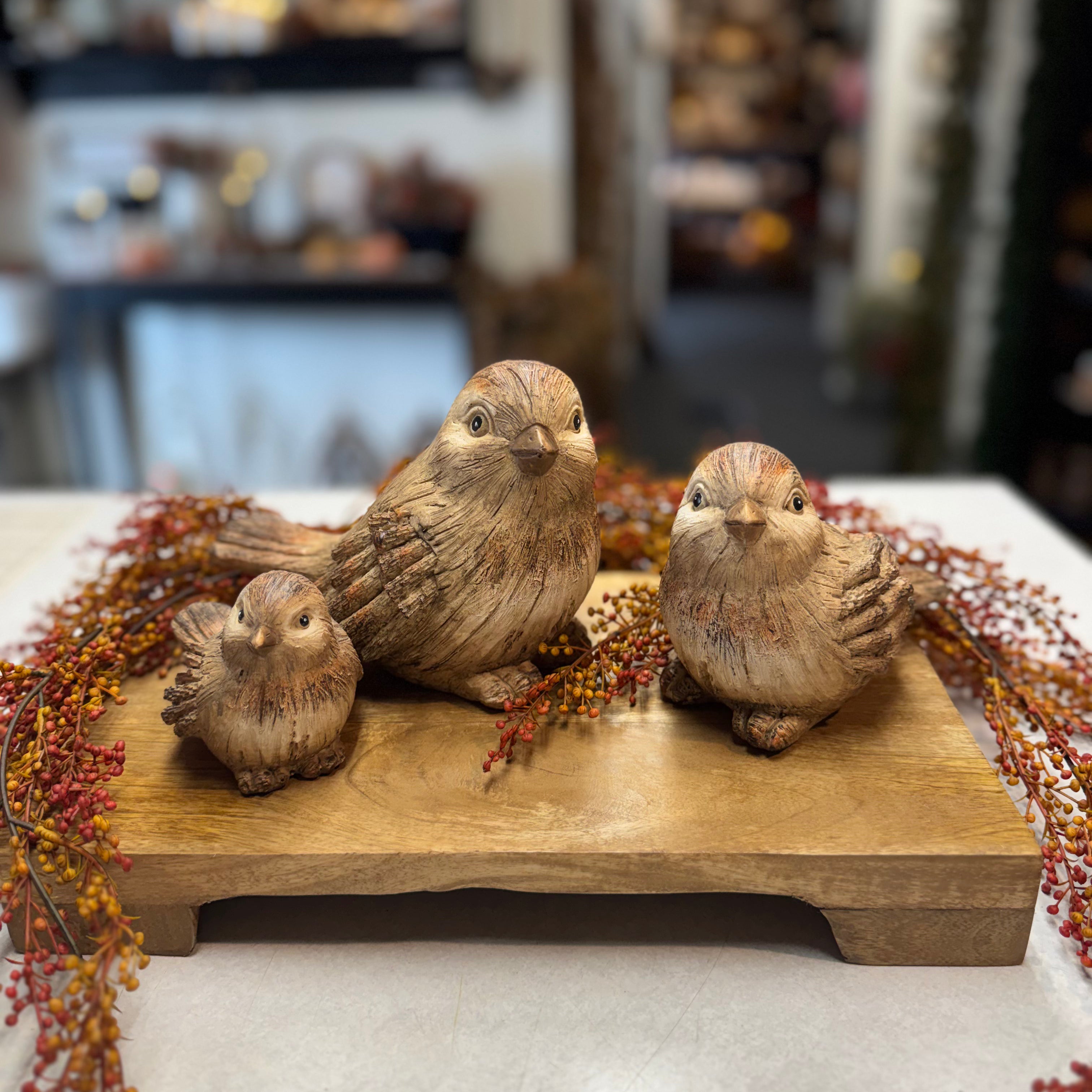 Three hand-carved wooden bird figurines in natural brown tones, arranged on a rectangular wooden base with dried autumn berries in a blurred indoor setting.