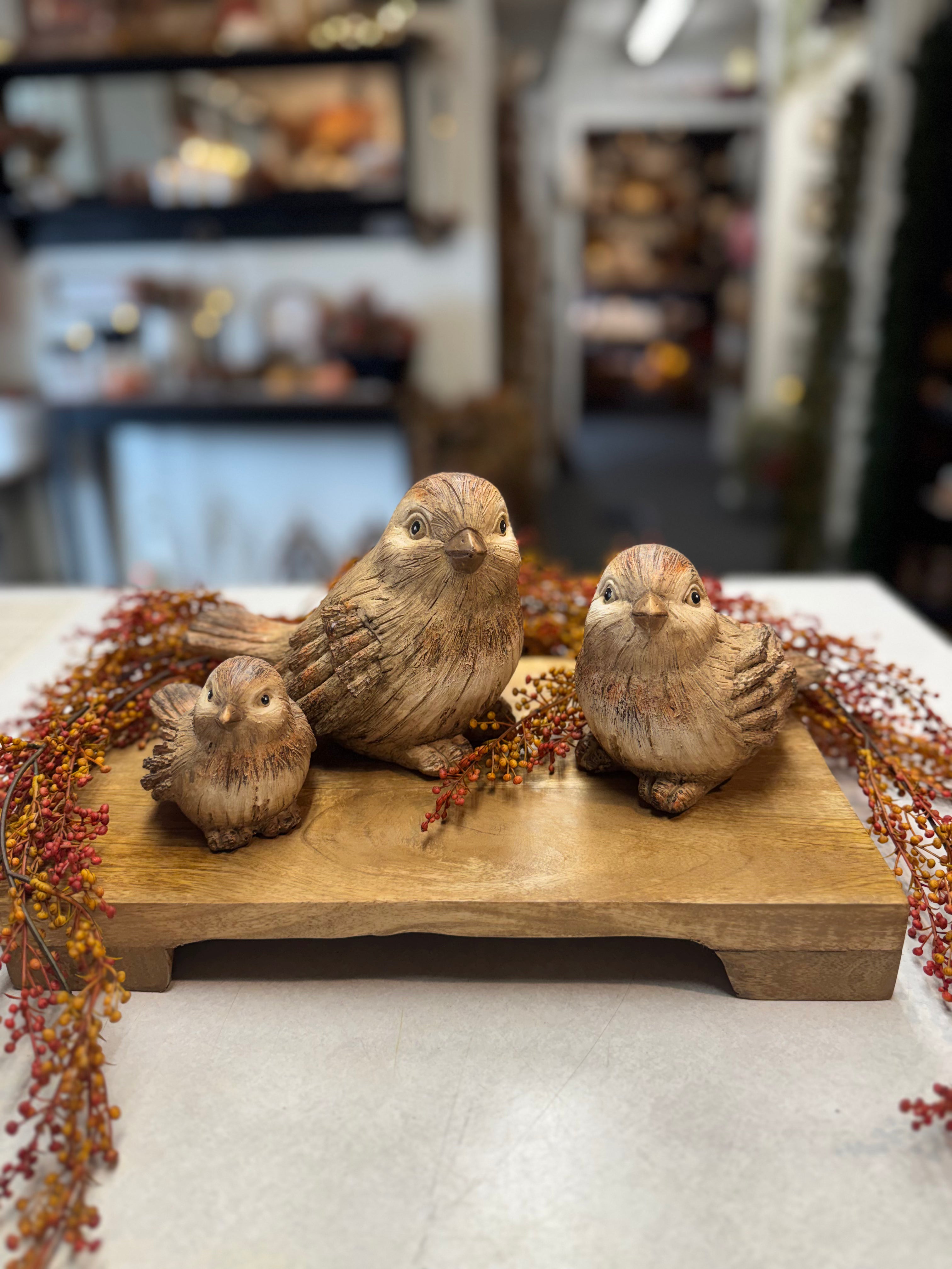 Three hand-carved wooden bird figurines in natural brown tones, arranged on a rectangular wooden base with dried autumn berries in a blurred indoor setting.