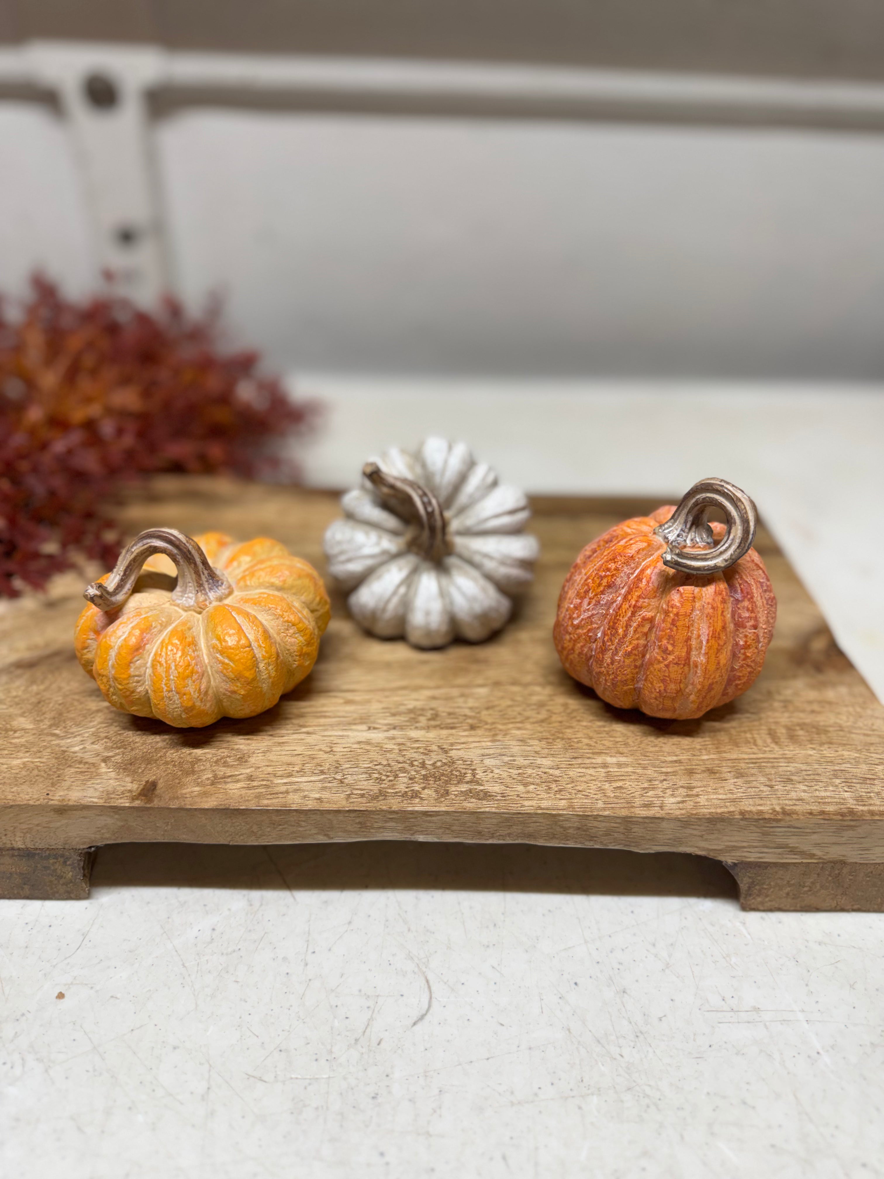 Three decorative miniature pumpkins in orange, white, and orange tones arranged on a wooden board, with a blurred neutral background.